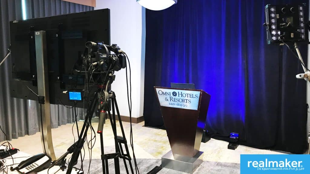 Ein Veranstaltungsraum mit einem Kamera-Setup und einem Podium mit dem Schild 'Omni Hotels & Resorts San Diego'. Im Hintergrund ist ein dunkelblauer Vorhang zu sehen. Links steht eine große Kamera auf einem Stativ, rechts befindet sich ein Lichtequipment. In der unteren Ecke ist ein Logo mit der Aufschrift 'realmaker Die Kreativagentur für Film und Web'.