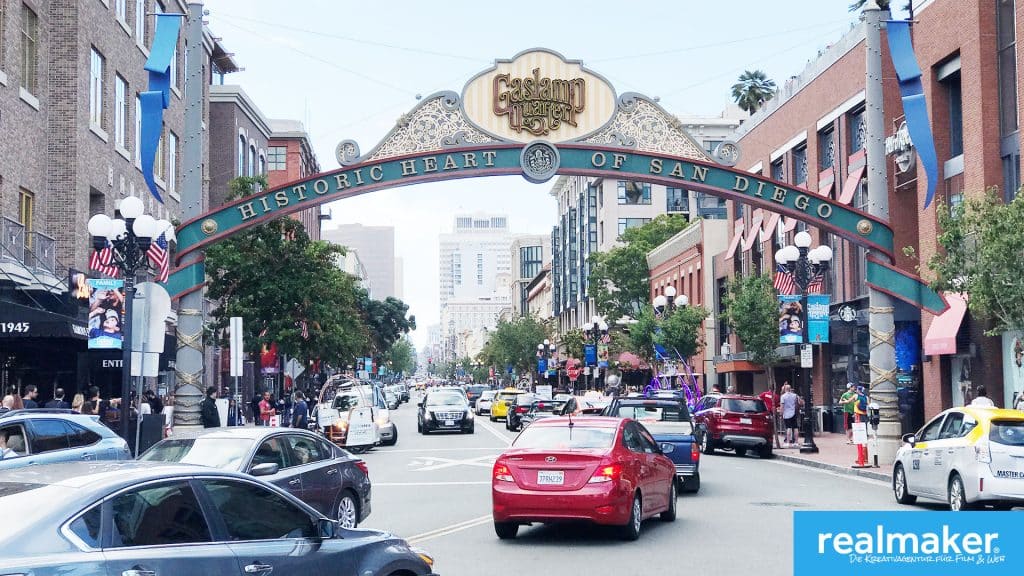 Das Bild zeigt das historische Gaslamp Quarter in San Diego. Über der Straße erstreckt sich ein verzierter Bogen mit der Aufschrift 'Gaslamp Quarter' und 'Historic Heart of San Diego'. Umgeben von lebendigen Straßen mit Autos und Fußgängern, ist der Blick von modernen bis hin zu historischen Gebäuden gesäumt. Im Vordergrund sind mehrere Autos sichtbar, darunter ein rotes Auto und ein Taxi. Auf der rechten unteren Ecke des Bildes befindet sich das Logo von realmaker, einer Kreativagentur für Film und Web.