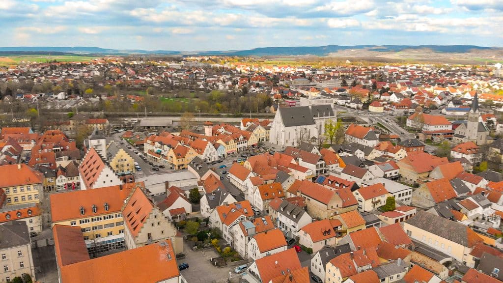 Luftaufnahme einer Stadt mit zahlreichen Gebäuden, die größtenteils rote Dächer haben. Im Zentrum steht eine große Kirche mit einem spitzen Turm. Die Umgebung ist ländlich mit Feldern und Hügeln am Horizont. Der Himmel ist bewölkt, aber es gibt auch einige sonnige Abschnitte.